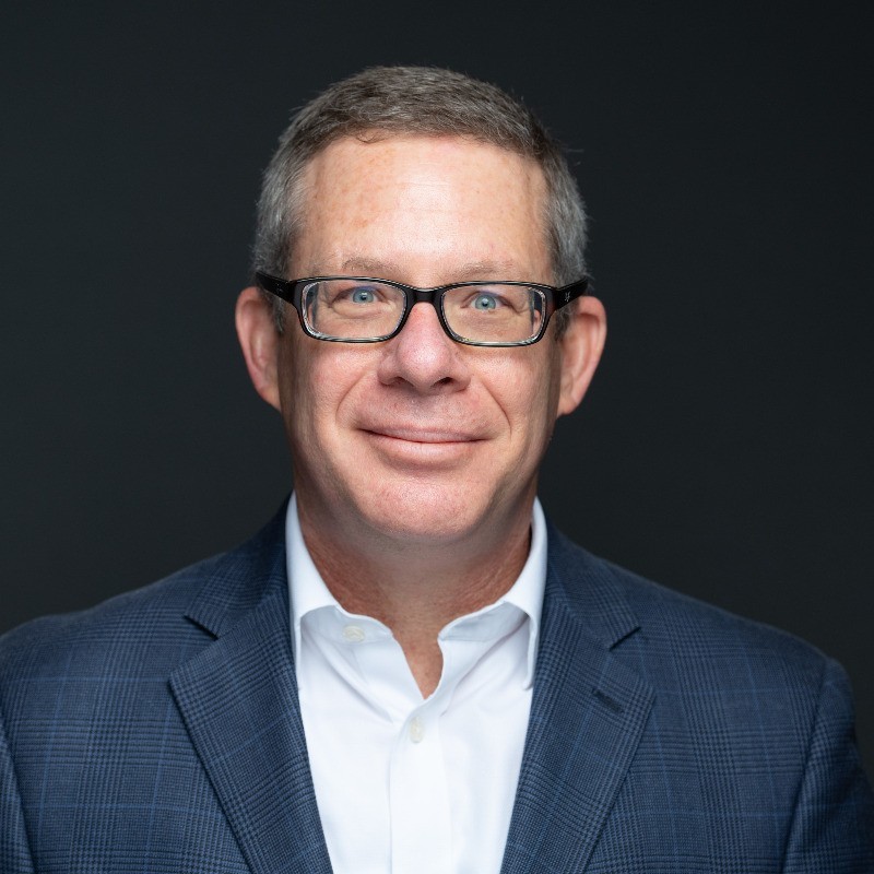 An image of Chris Mostek, a cleanshaven man with short hair and glasses wearing a suit.