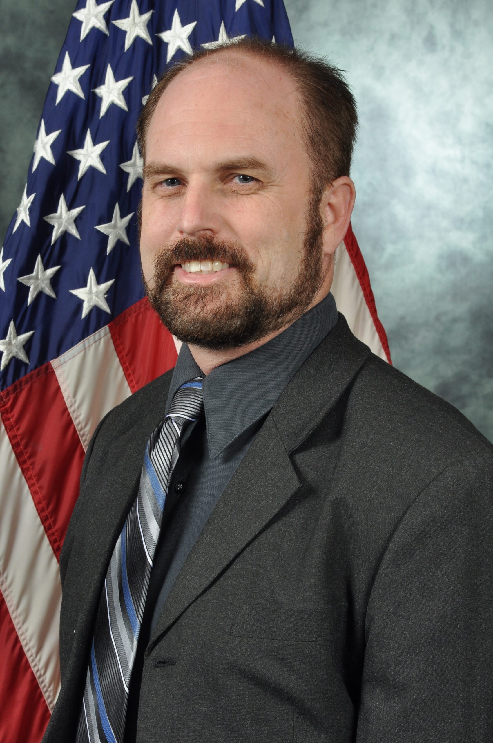 A photo of a smiling white man with dark hair, beard, and moustache wearing a dark suit and standing in front of a US flag.