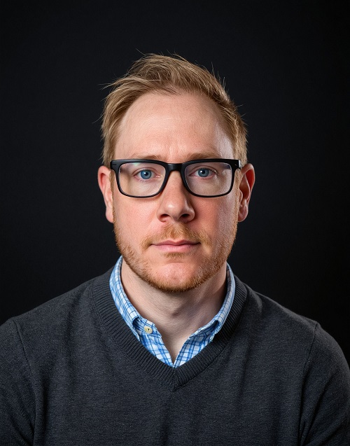 An AI-edited photo of RSVP Board Member Jay Sherrill. A man wearing a blue shirt and grey sweater and with short hair, a little bit of facial hair, and dark-rimmed glasses.